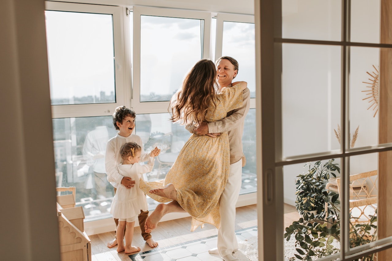 Na zdjęciu widać szczęśliwą rodzinę, składającą się z rodziców i dwójki dzieci, spędzających czas razem w sypialni. Rodzice uśmiechają się i przytulają. Dzieci stoją radośnie przy dużym oknie.